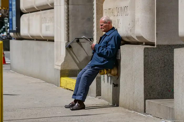 an older man reads a newspaper leaning against a bank